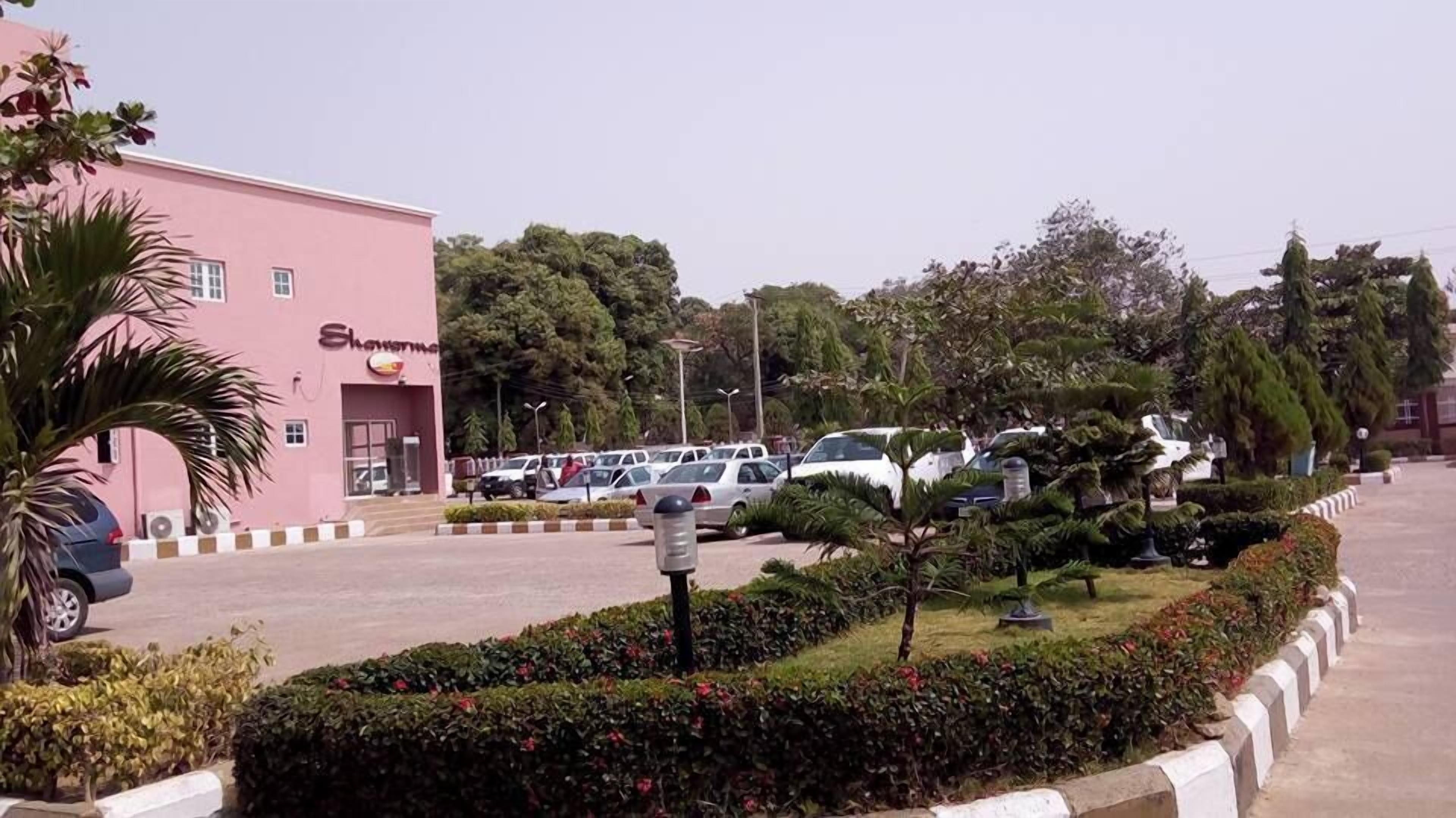 Ein pinkfarbenes Gebäude mit einem Schild, auf dem "Showroom" steht. Vor dem Gebäude sind Autos geparkt, und ein gepflegter Garten mit Hecken und einem Laternenpfahl.