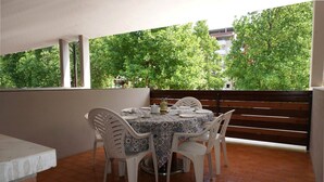 En terrasse med et rundt bord dækket til fire personer, omgivet af hvide stole, med flisegulv og en træbænk i baggrunden.