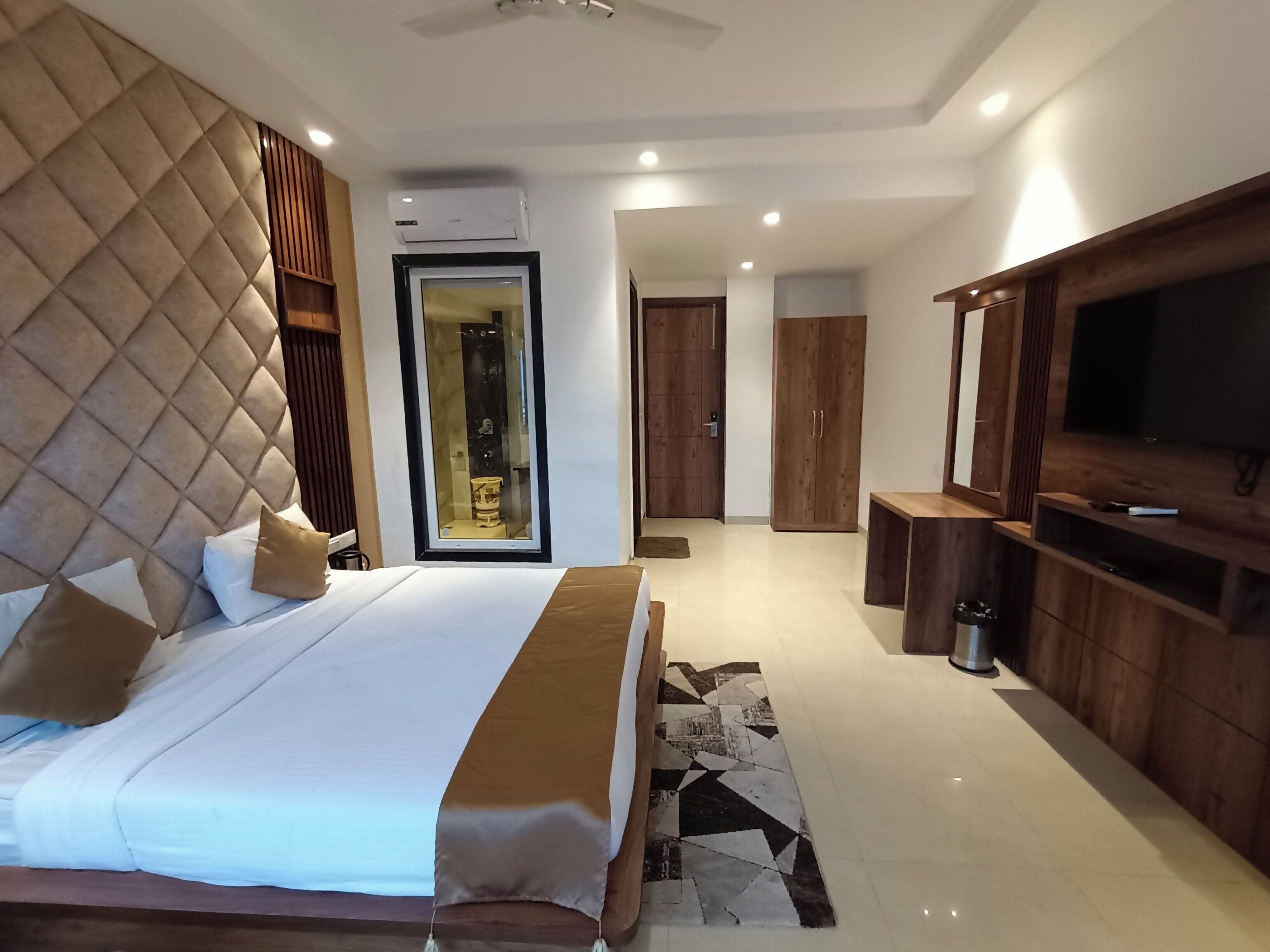 Quarto de hotel moderno com uma cama grande, televisão de tela plana, móveis de madeira e banheiro visível por uma porta de vidro.