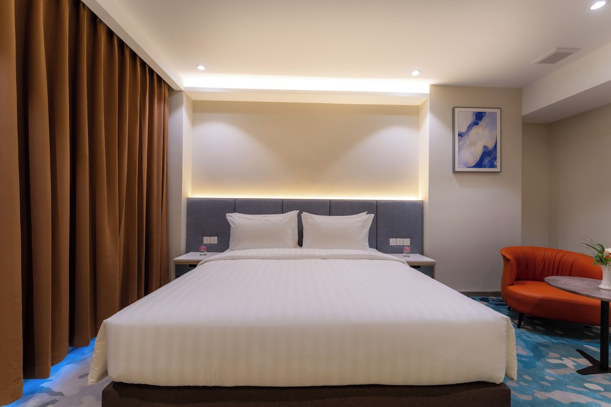Habitación de hotel con una cama grande, una mesita de noche con lámpara, una silla naranja y una pequeña mesa con un jarrón.