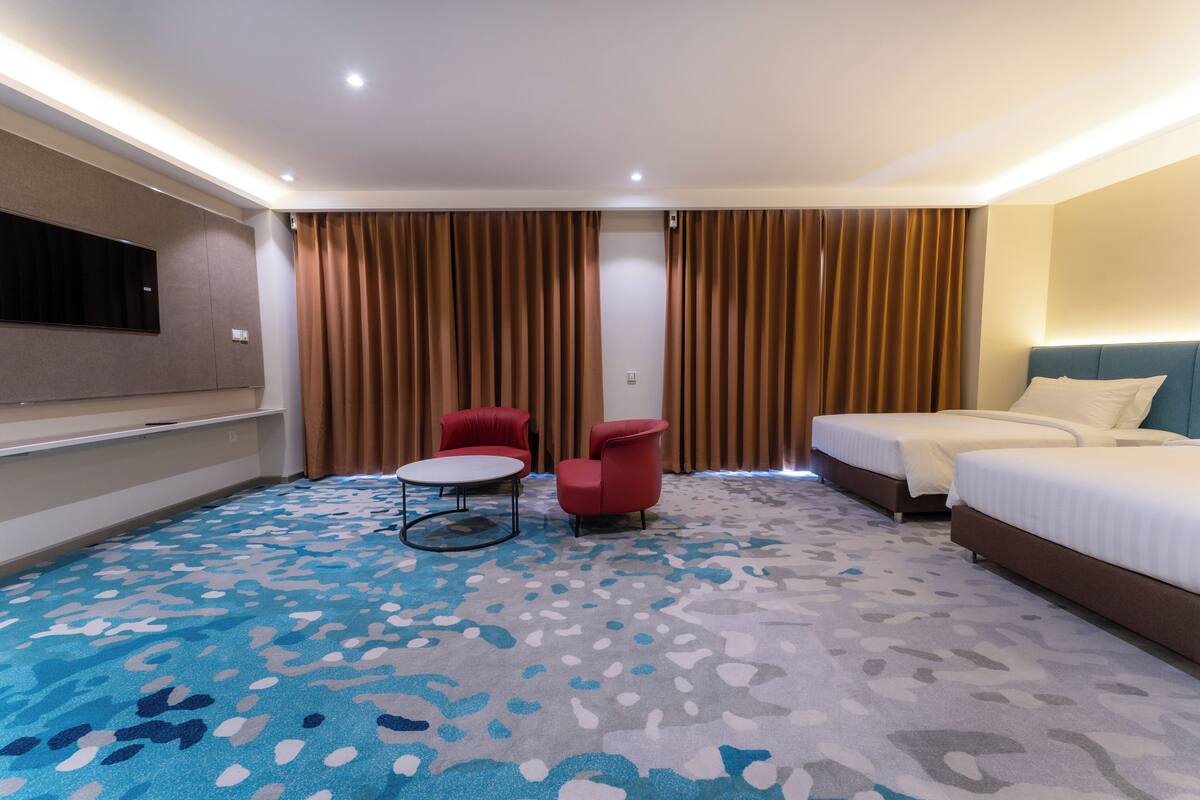 Ein Hotelzimmer mit einem großen Bett, einem kleineren Bett, einem an der Wand befestigten Fernseher, einem runden Tisch, zwei roten Stühlen und einem gemusterten Teppich.