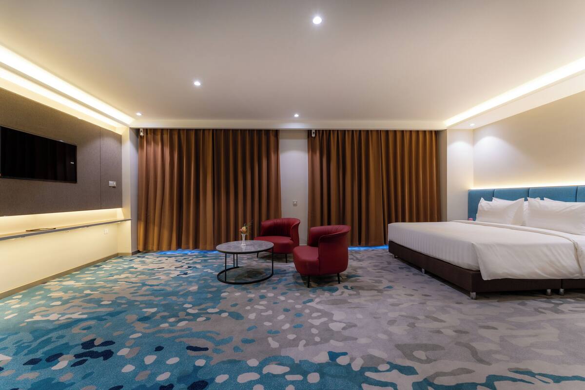 Ein Hotelzimmer mit einem großen Bett, zwei roten Sesseln, einem kleinen runden Tisch und einem an der Wand befestigten Fernseher.