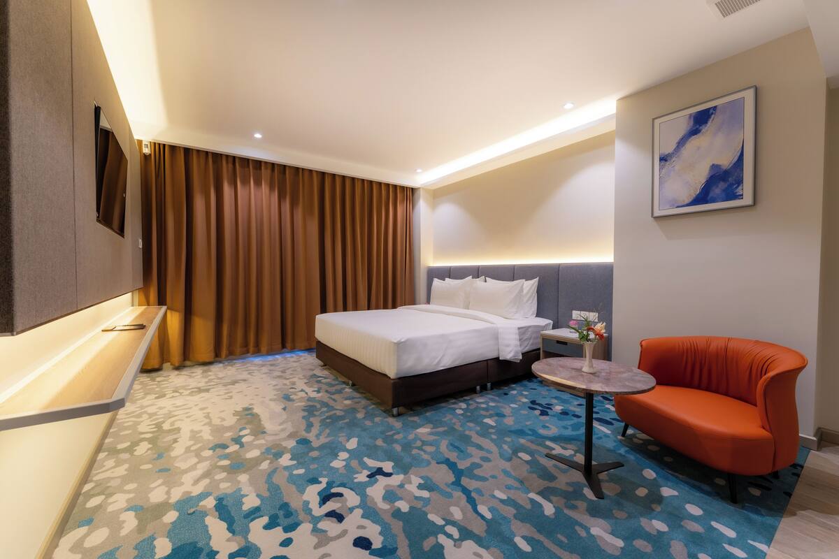 Ein Hotelzimmer mit einem Bett, einem kleinen runden Tisch, einem orangefarbenen Sessel, einem Bild an der Wand und einem gemusterten Teppich.