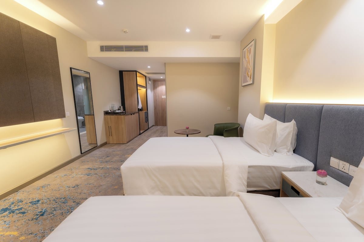 Ein Hotelzimmer mit zwei Betten, einem kleinen Tisch, einem grünen Sessel und einer Küchenzeile im Hintergrund.