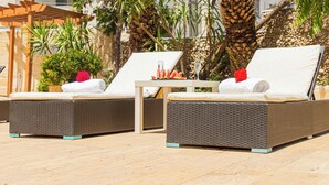 Dos sillones de mimbre con cojines blancos, una mesita con un vaso de agua y una fuente de fruta, y un fondo con palmeras y un edificio.