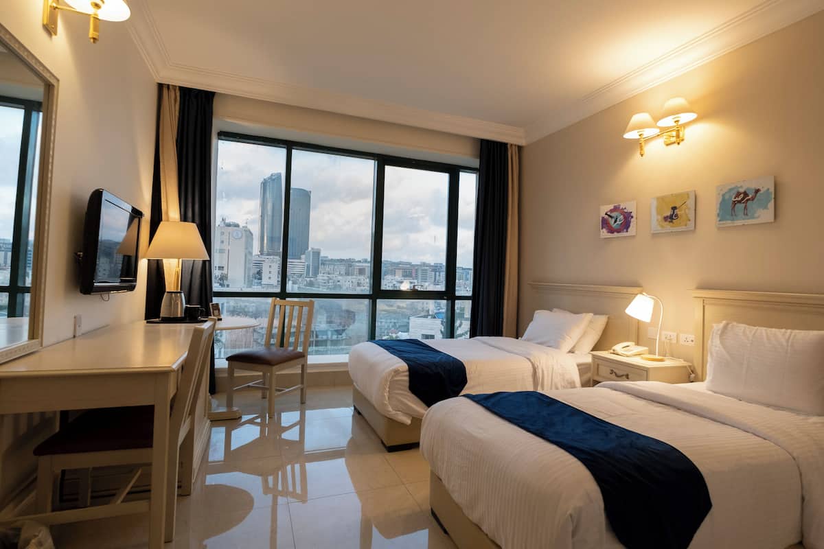 Habitación de hotel con dos camas, un escritorio, una silla, un televisor y un amplio ventanal con vistas a la ciudad.