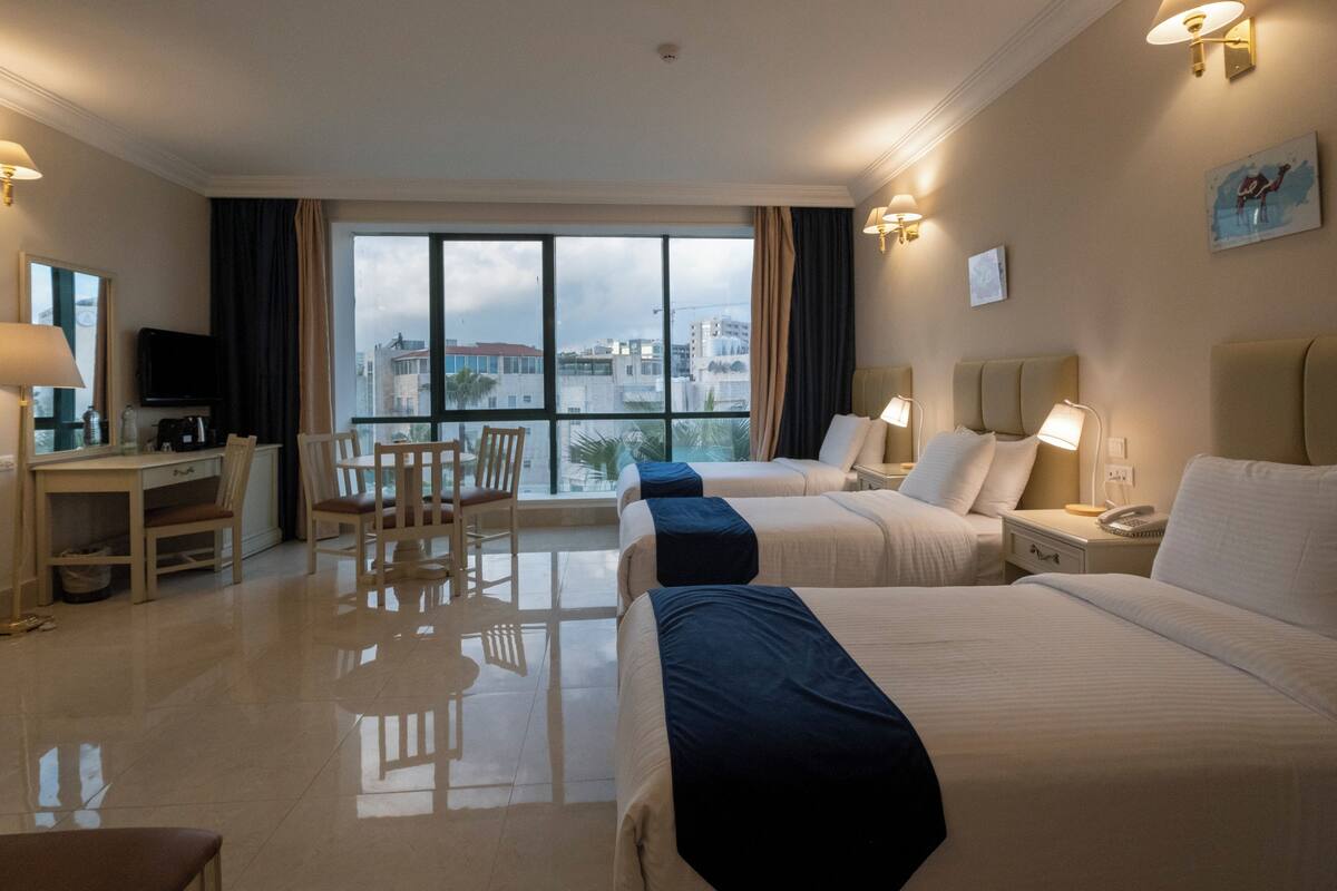 Habitación de hotel con dos camas, un escritorio, una silla y un amplio ventanal con vistas a la ciudad.