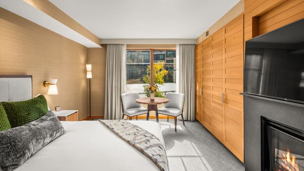 A hotel room with a bed, a fireplace, a seating area with two chairs, a table, and a window with curtains. - Luxury Resort Ski In Ski Out Studio (Snowmass Village)
