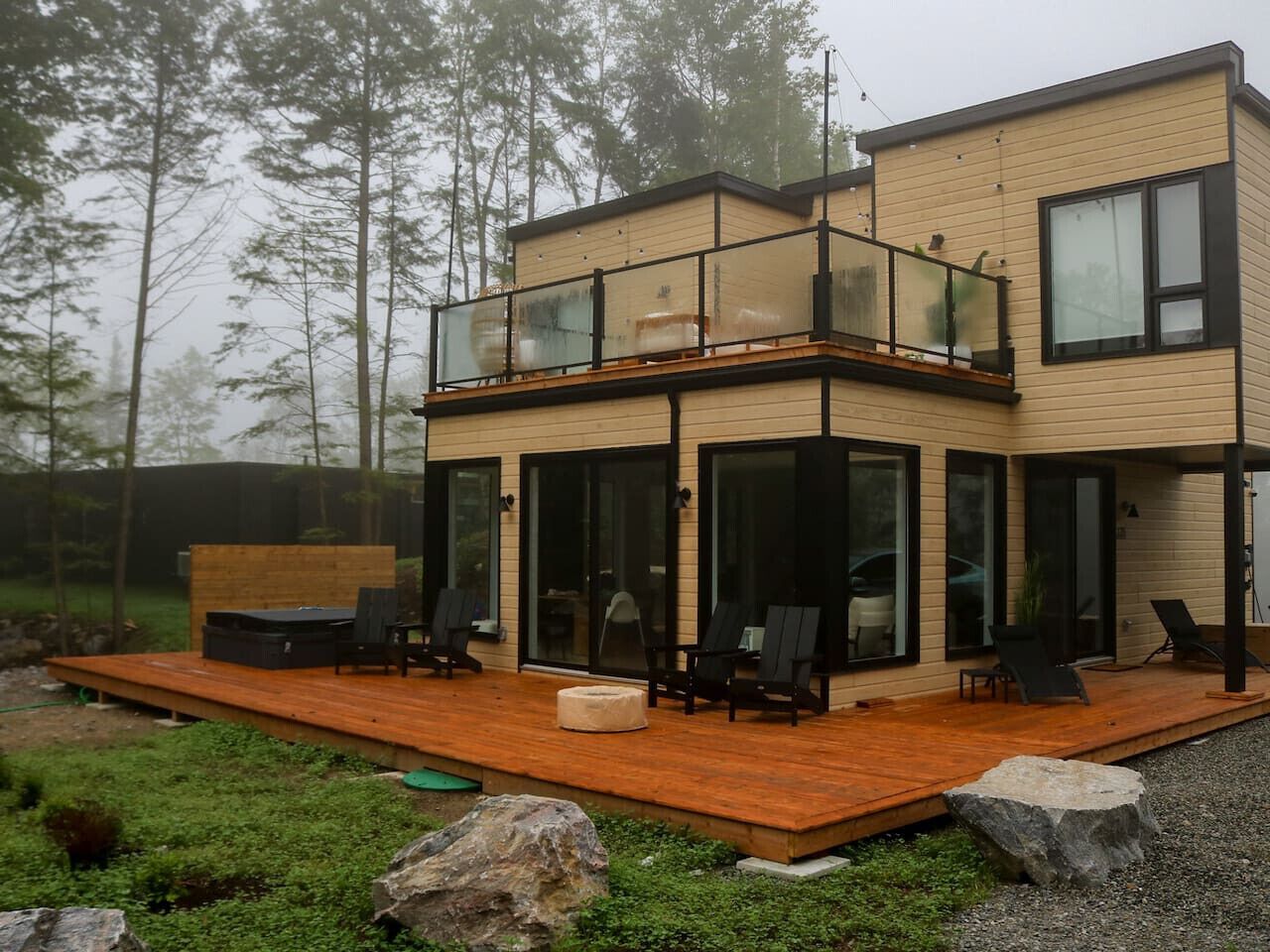 Ein modernes, zweistöckiges Haus mit einer Holzdachterrasse, Glasgeländern und Sitzgelegenheiten im Freien.