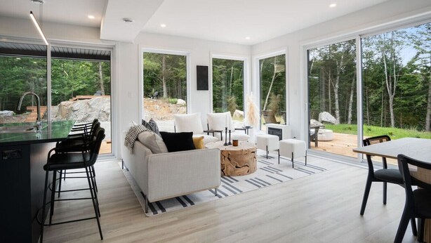 A modern interior with a kitchen, dining area, and seating by a large window overlooking a natural landscape.