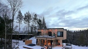 A modern two-story house with a deck, surrounded by snow and trees.