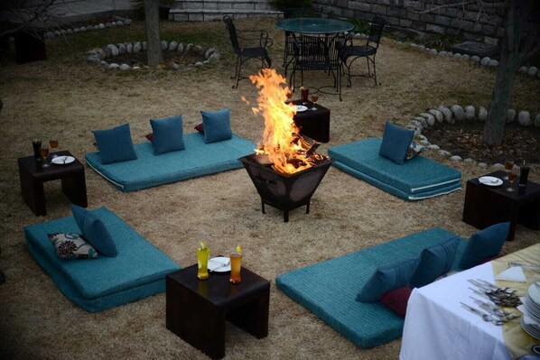 An outdoor seating area with blue cushioned sofas, a fire pit, and a table set for dining. - Mandawara - Shri Orchard House (Kullu)