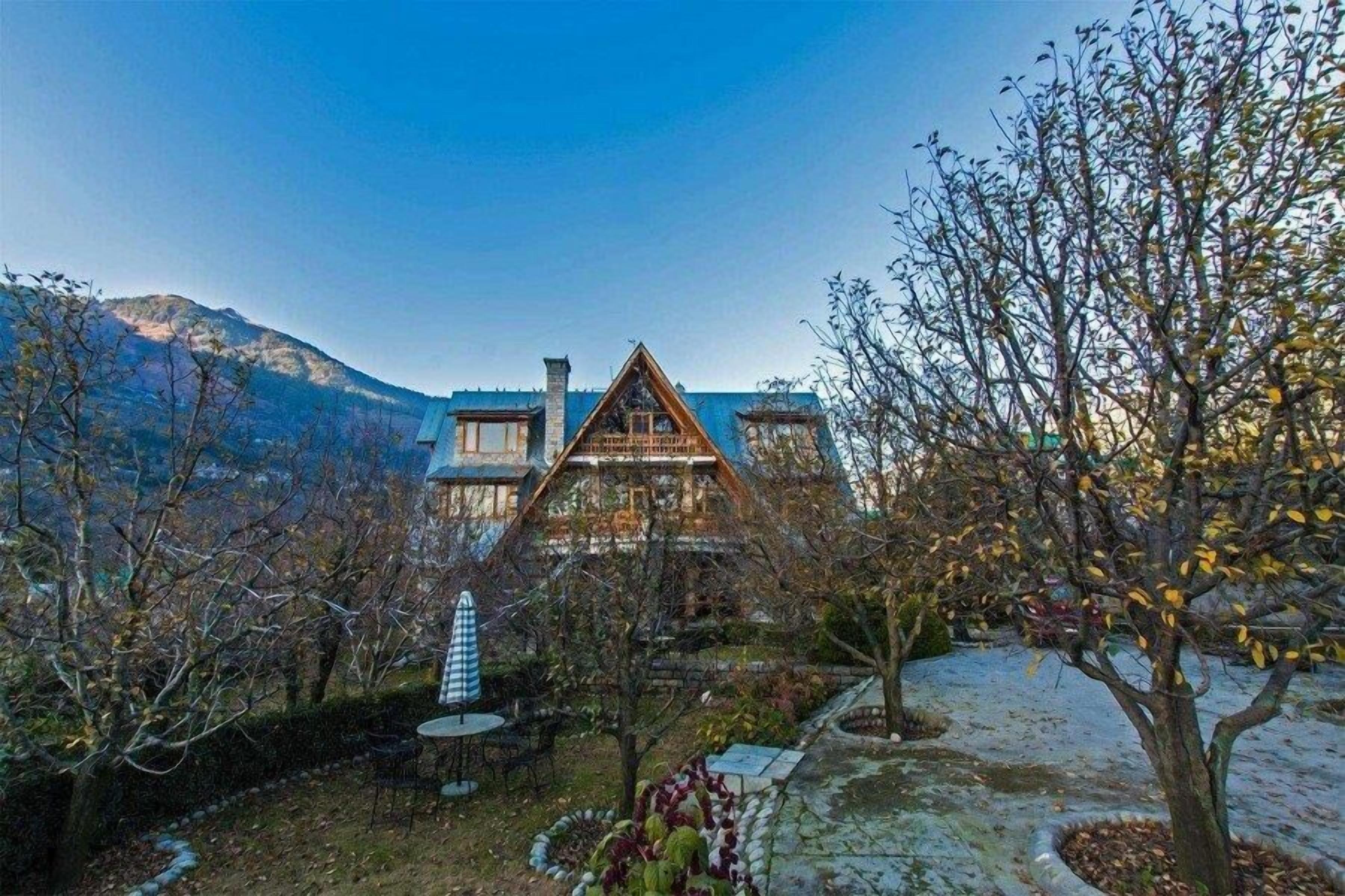 A chalet-style house with a steeply pitched roof, surrounded by a garden with a variety of plants and trees.