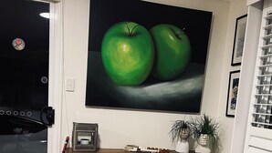 Un comedor con una mesa de madera, un cuenco de frutas, un cuadro con manzanas y una pequeña estantería con objetos decorativos.