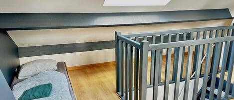 A room with a sloped ceiling, a wooden floor, a bed with a patterned bedspread, a grey crib, and a skylight.