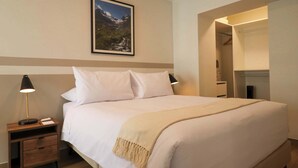 Een net opgemaakt bed met witte lakens, een beige plaid en een nachtkastje met een lamp en een boek.