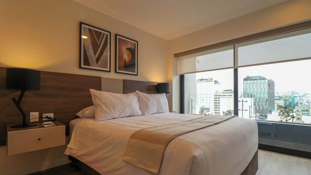 A modern hotel room with a large bed, a nightstand, a lamp, and a view of the city through large windows. - w* | Comfy 2BR w/ Balcony Near Larcomar (Miraflores)