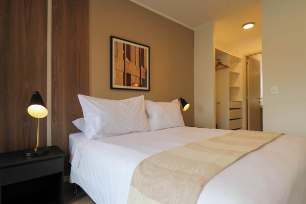 A neatly made bed with white linens and a beige throw blanket, two bedside lamps, a framed abstract artwork on the wall, and a wooden wardrobe in the background.