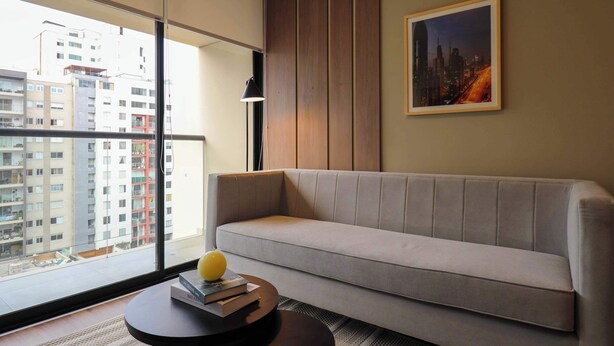 Ein modernes Wohnzimmer mit einer Couch, einem runden Couchtisch mit Büchern und einem gelben Gegenstand, einem gerahmten Bild an der Wand und Blick auf Gebäude durch ein großes Fenster.
