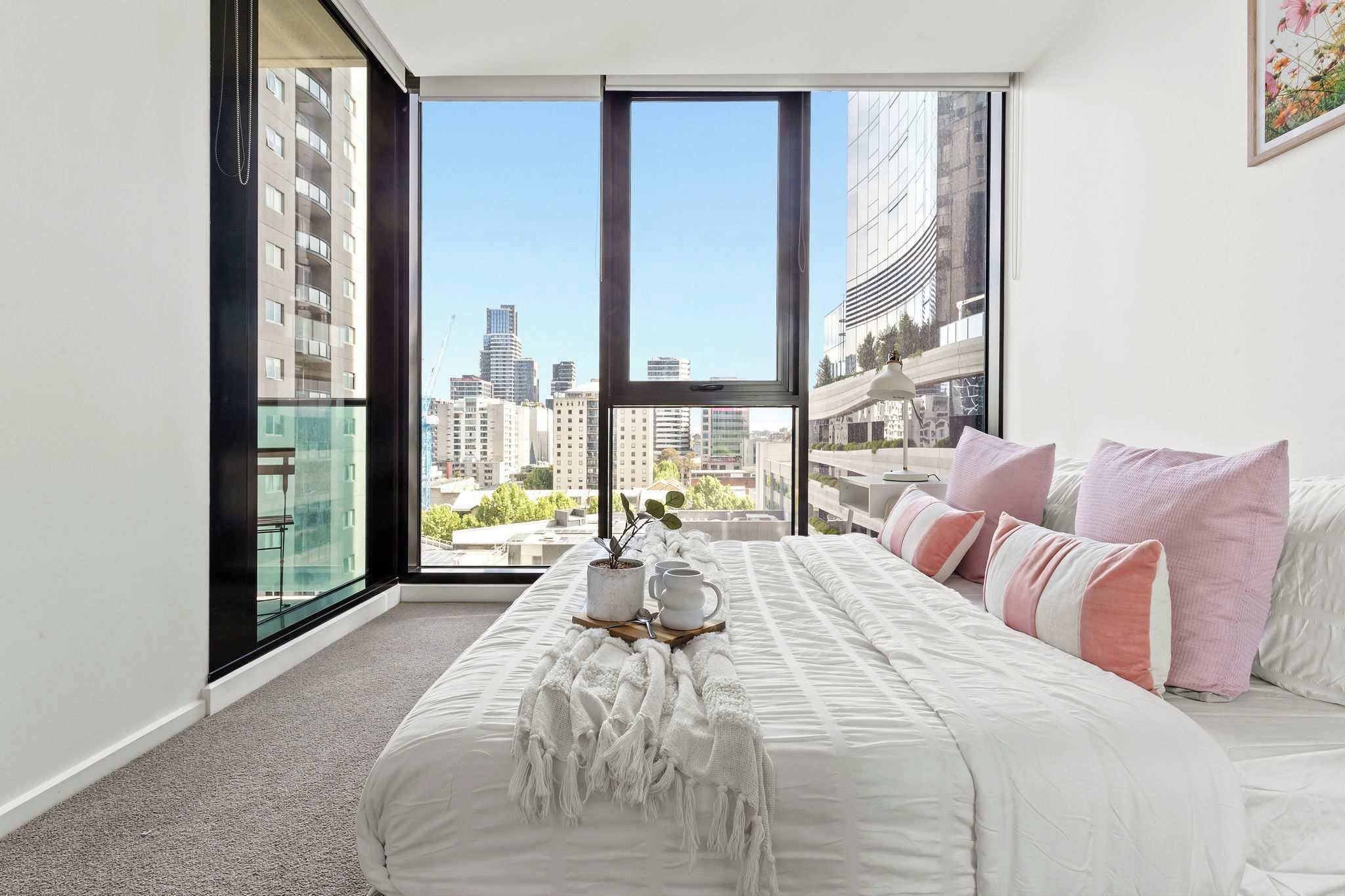 A bedroom with a large window, a bed with white bedding, and a view of the cityscape.