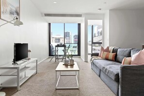 Een moderne woonkamer met een grijze bank, een flatscreen televisie op een witte standaard, een klein witte salontafel, en een balkon met uitzicht op gebouwen.