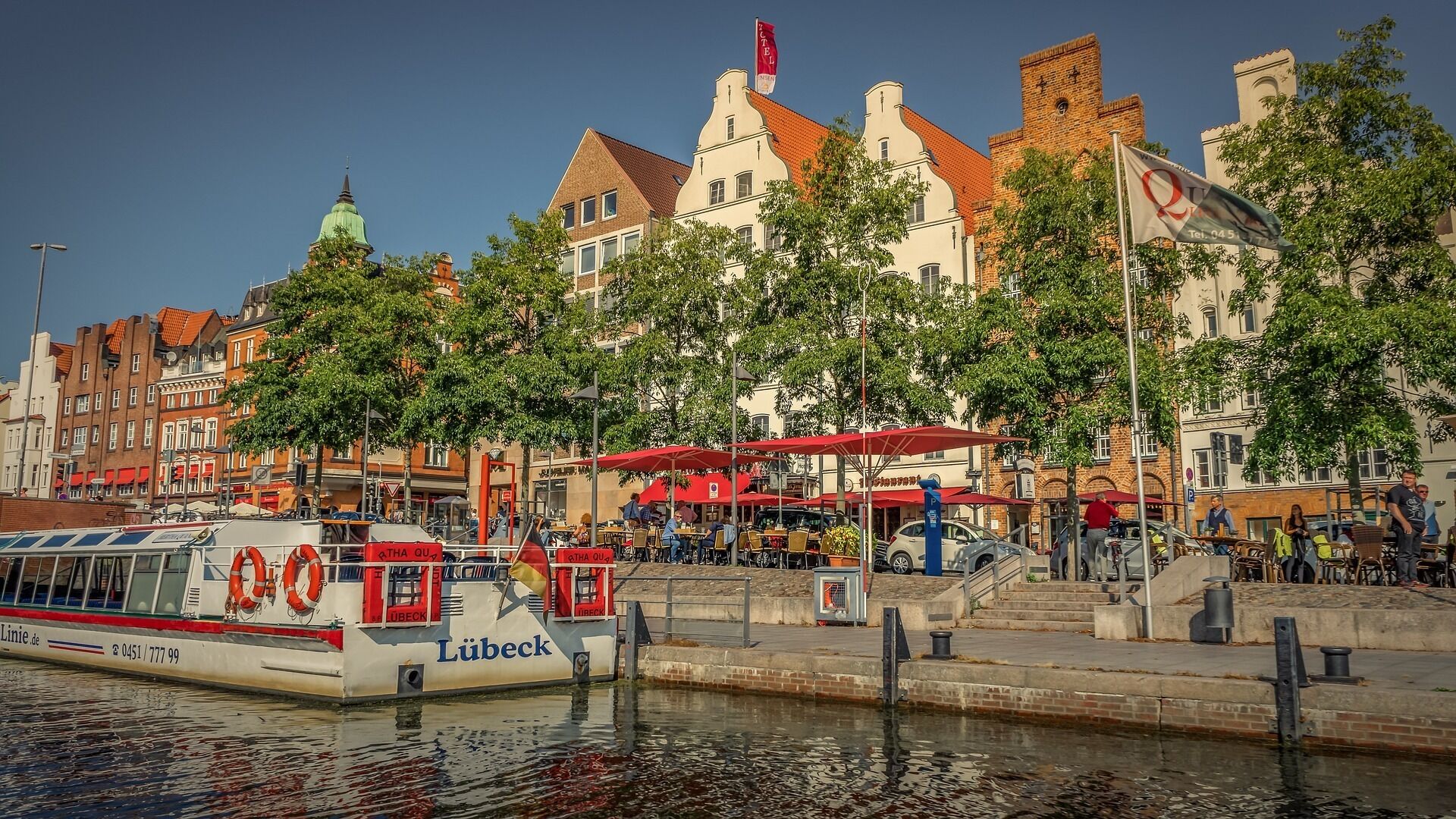 Ein Boot namens 'Lübeck' liegt an einem Kai, im Hintergrund eine Reihung von Gebäuden.