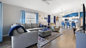 Un soggiorno moderno con un divano angolare grigio, una poltrona blu e un poggiapiedi. Si vede una ventola a soffitto, un'ampia finestra con tende blu, e un angolo cucina sullo sfondo.