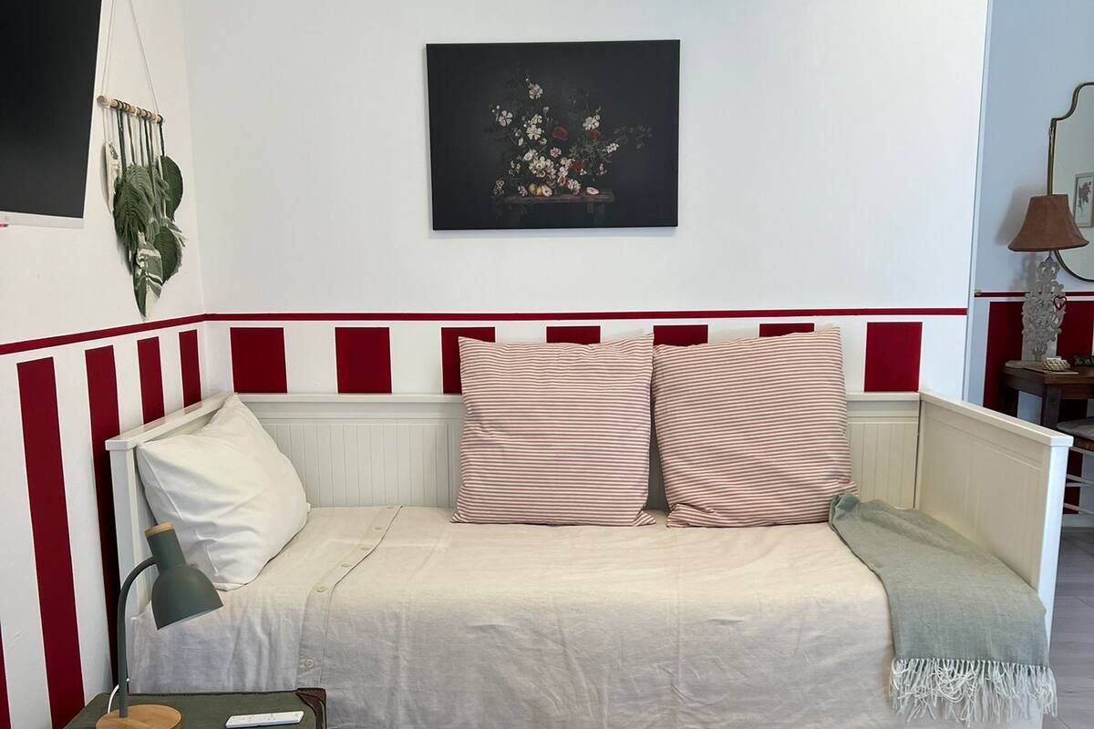 A cozy living room with a white sofa, striped pillows, a small table with a lamp, and a framed artwork on the wall.