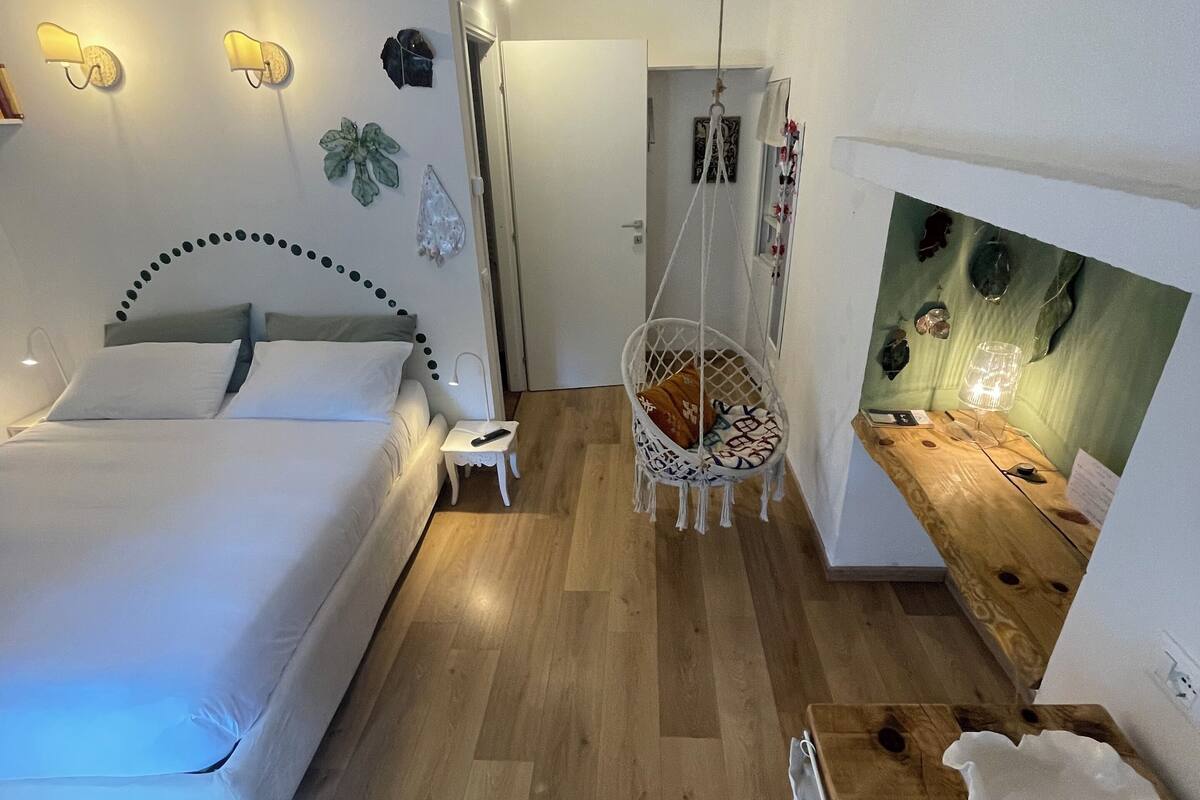 A bedroom with a bed, bedside table, hanging chair, and a wall-mounted light fixture.