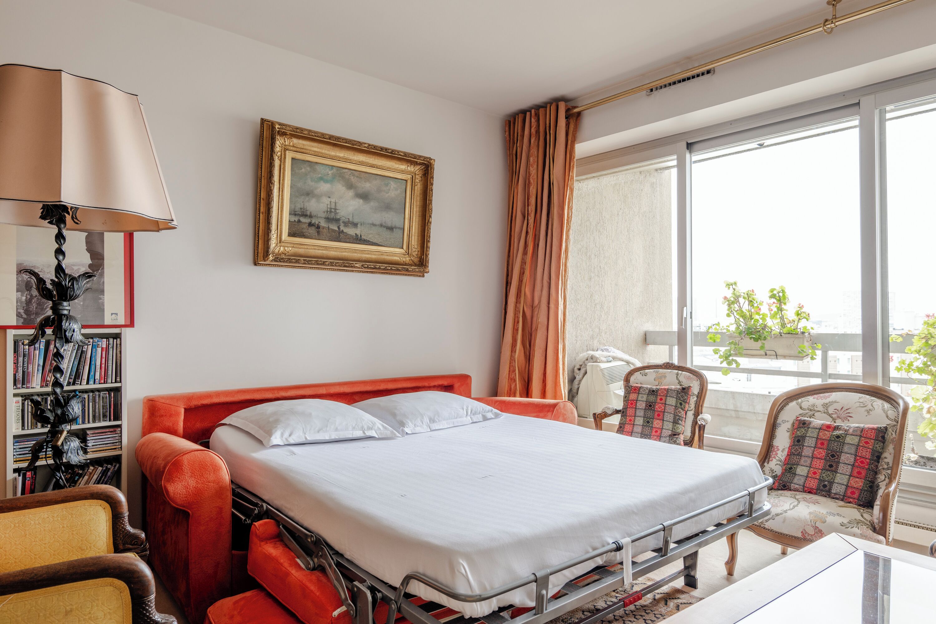 A hotel room with a bed, a bookshelf, a lamp, a chair, and a window with curtains.