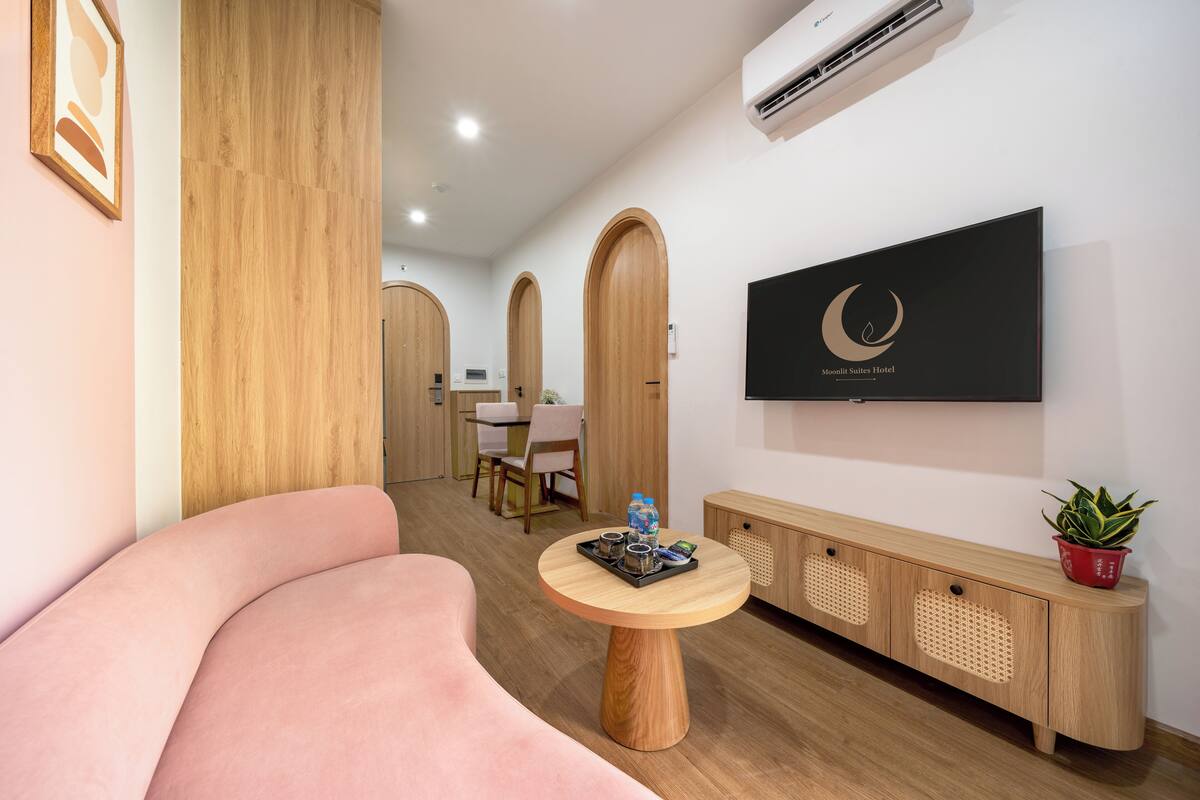 Un hall d’hôtel moderne avec un canapé rose aux lignes courbes, une console en bois agrémentée d’une plante et un téléviseur affichant le nom de l’hôtel.