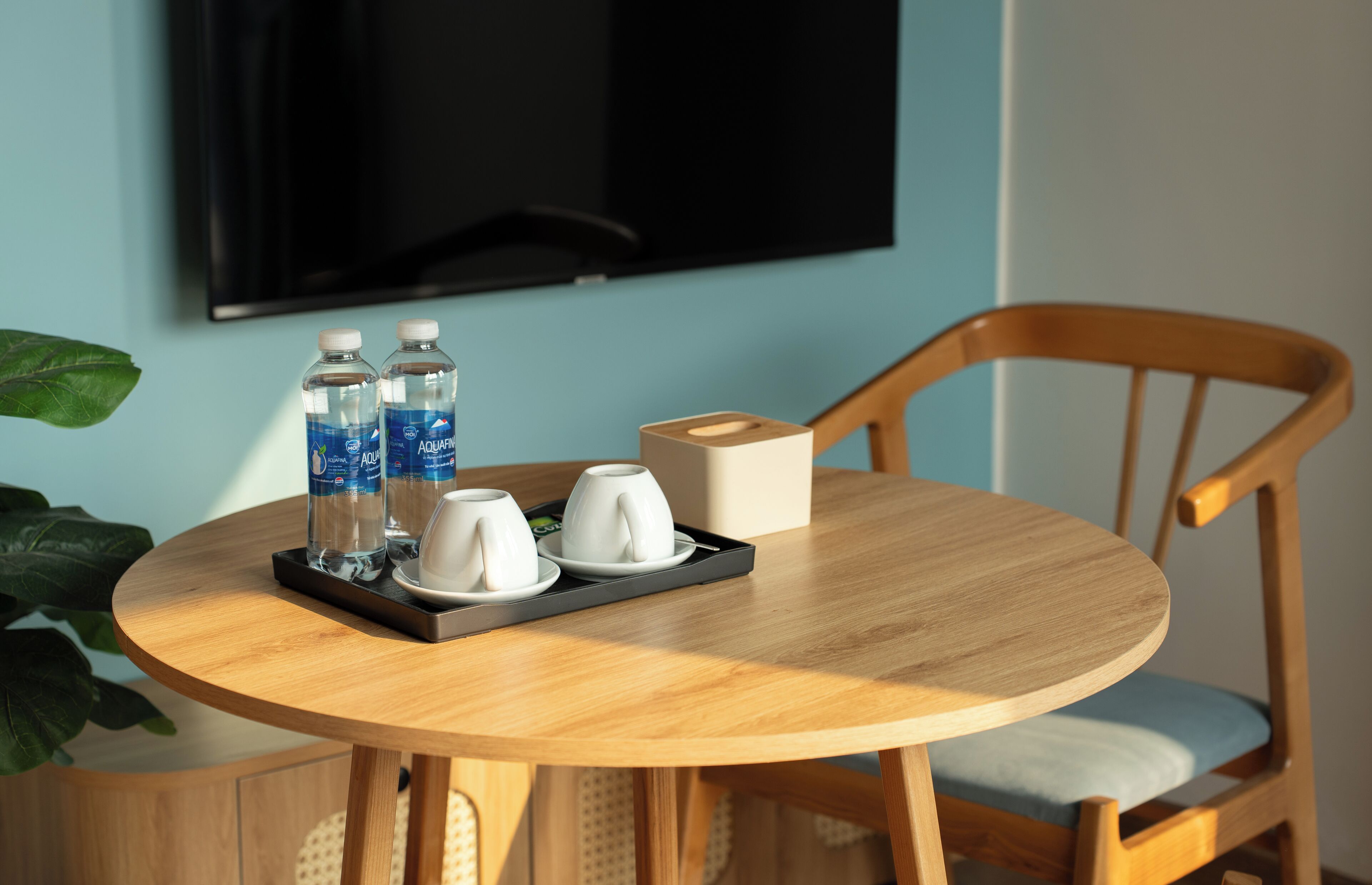 A round wooden table with a tray holding two water bottles, two white cups, and a tissue box. A wooden chair with a light blue cushion is positioned at the table.