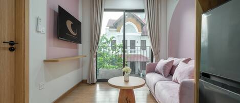 A modern living room with a round wooden coffee table, a pink sofa, and a large window with a view of a building and greenery outside.