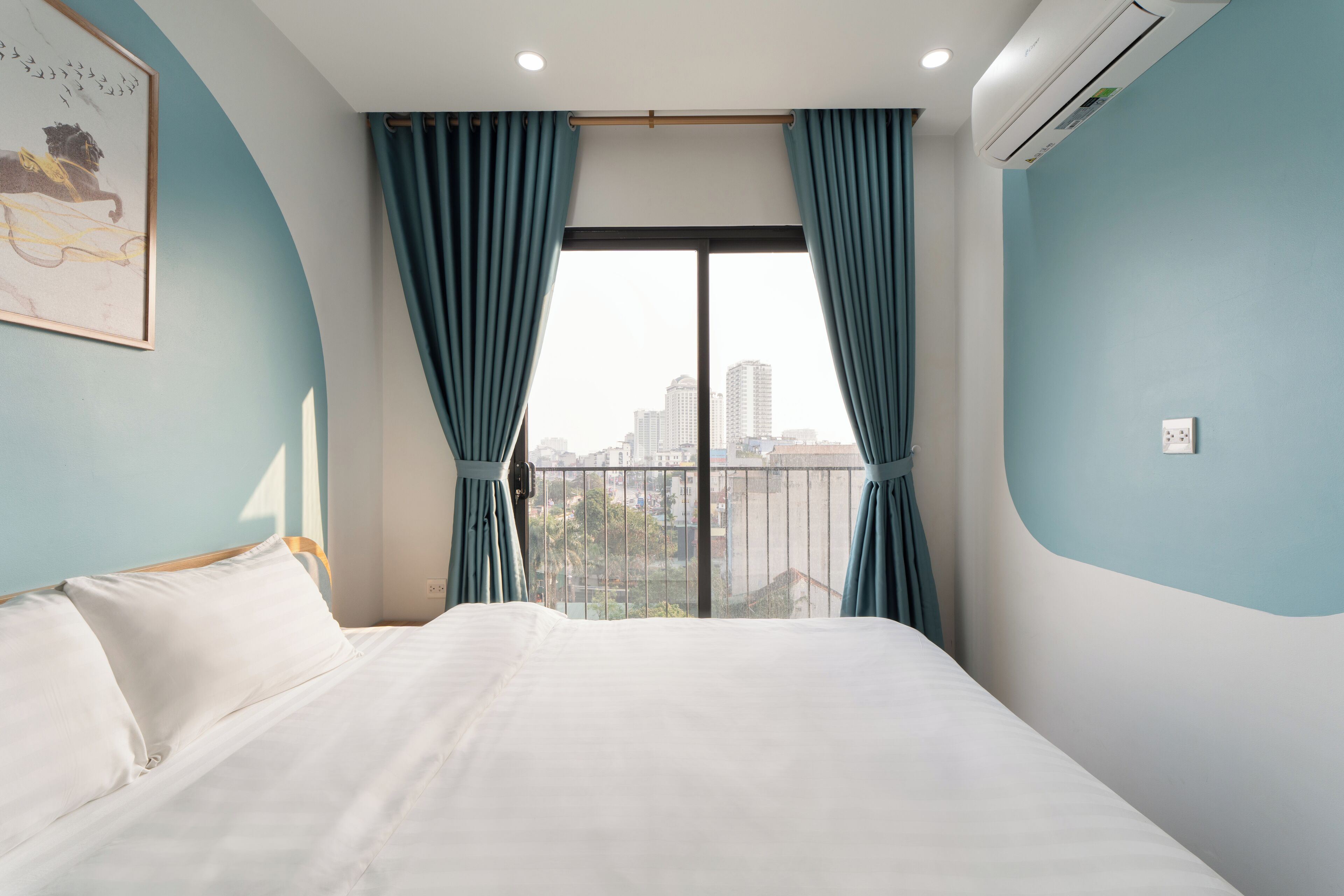 A hotel room with a bed, a window with curtains, a city view, and an air conditioning unit.