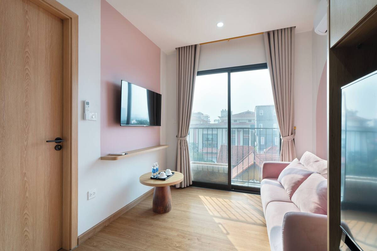 Une chambre d’hôtel moderne avec une porte en bois, un téléviseur à écran plat fixé au mur, une table ronde en bois avec un plateau, un canapé rose et une grande fenêtre avec des rideaux.