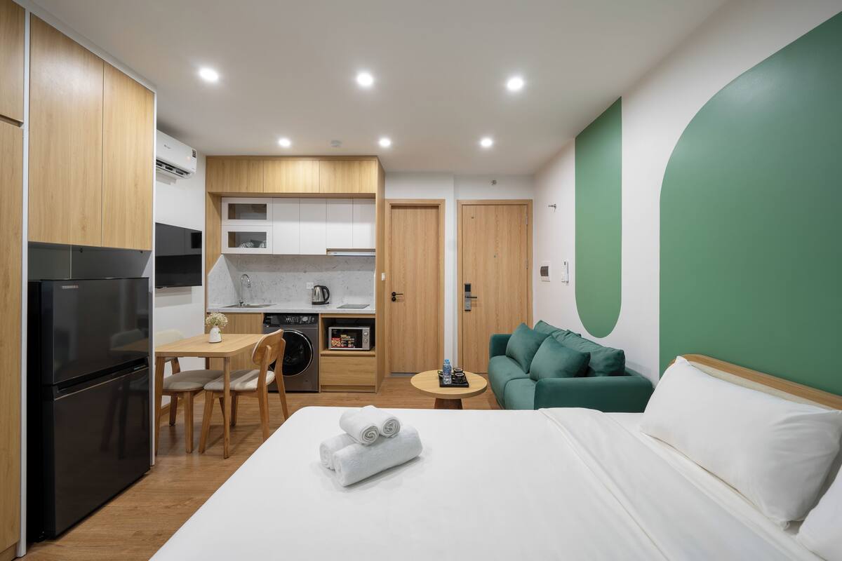 Une chambre d’hôtel compacte dotée d’une kitchenette, d’un lit avec du linge de lit blanc et d’un mur d’accent vert.