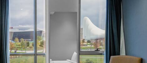 Hotelkamer met een groot bed, een bruine fauteuil, een klein tafeltje met een vaas en fruit, en uitzicht op een modern gebouw en groen.