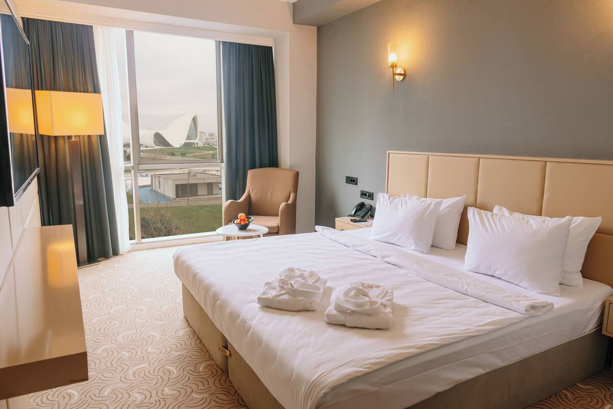 Ein Hotelzimmer mit einem großen Bett, einem Sessel, einem Schreibtisch und Blick auf ein Gebäude.