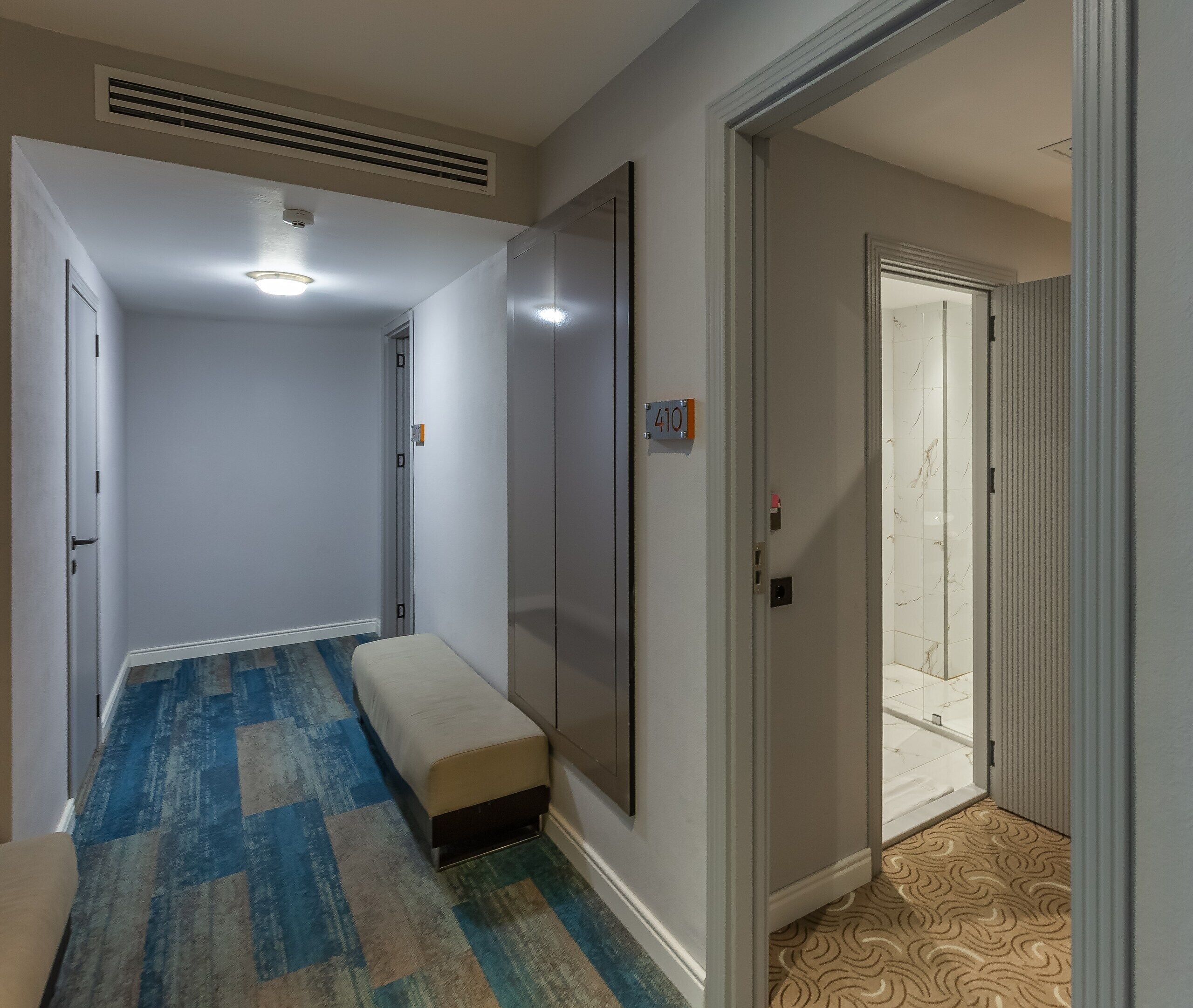 A hallway with a bench, a door with a number sign, and a patterned carpet.