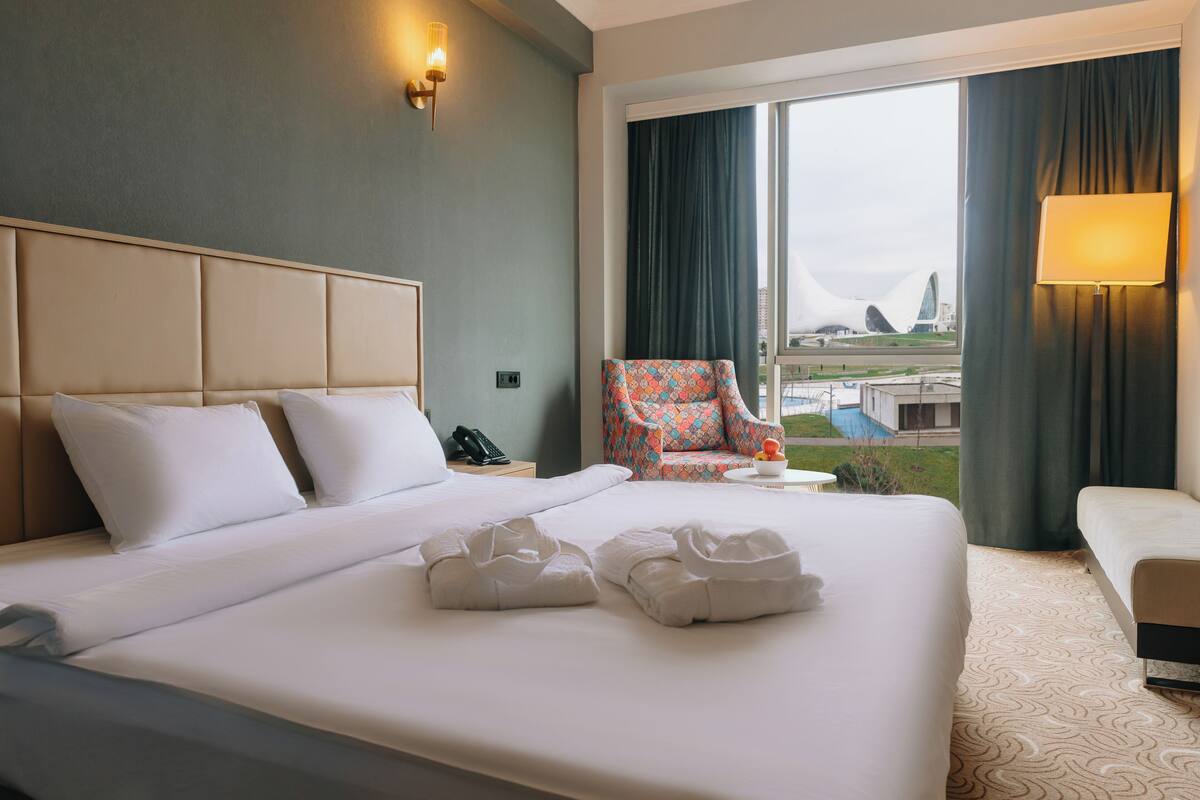 Ein Hotelzimmer mit einem großen Bett, einem Sessel, einer Lampe und Blick auf ein Gebäude.