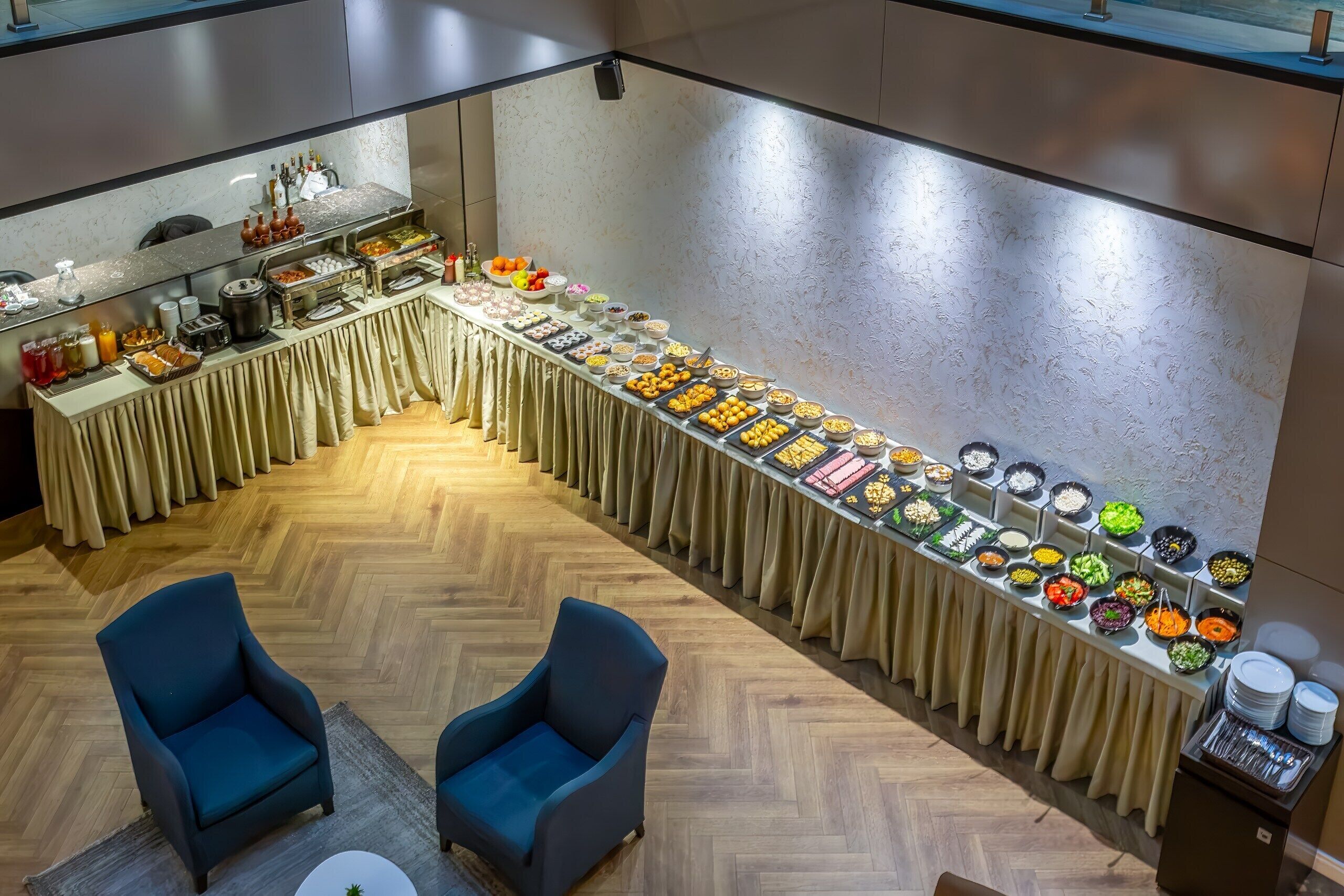 A buffet setup with various food items and beverages, two blue armchairs, and a wooden floor.