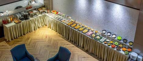 A buffet setup with various food items and beverages, two blue armchairs, and a wooden floor.