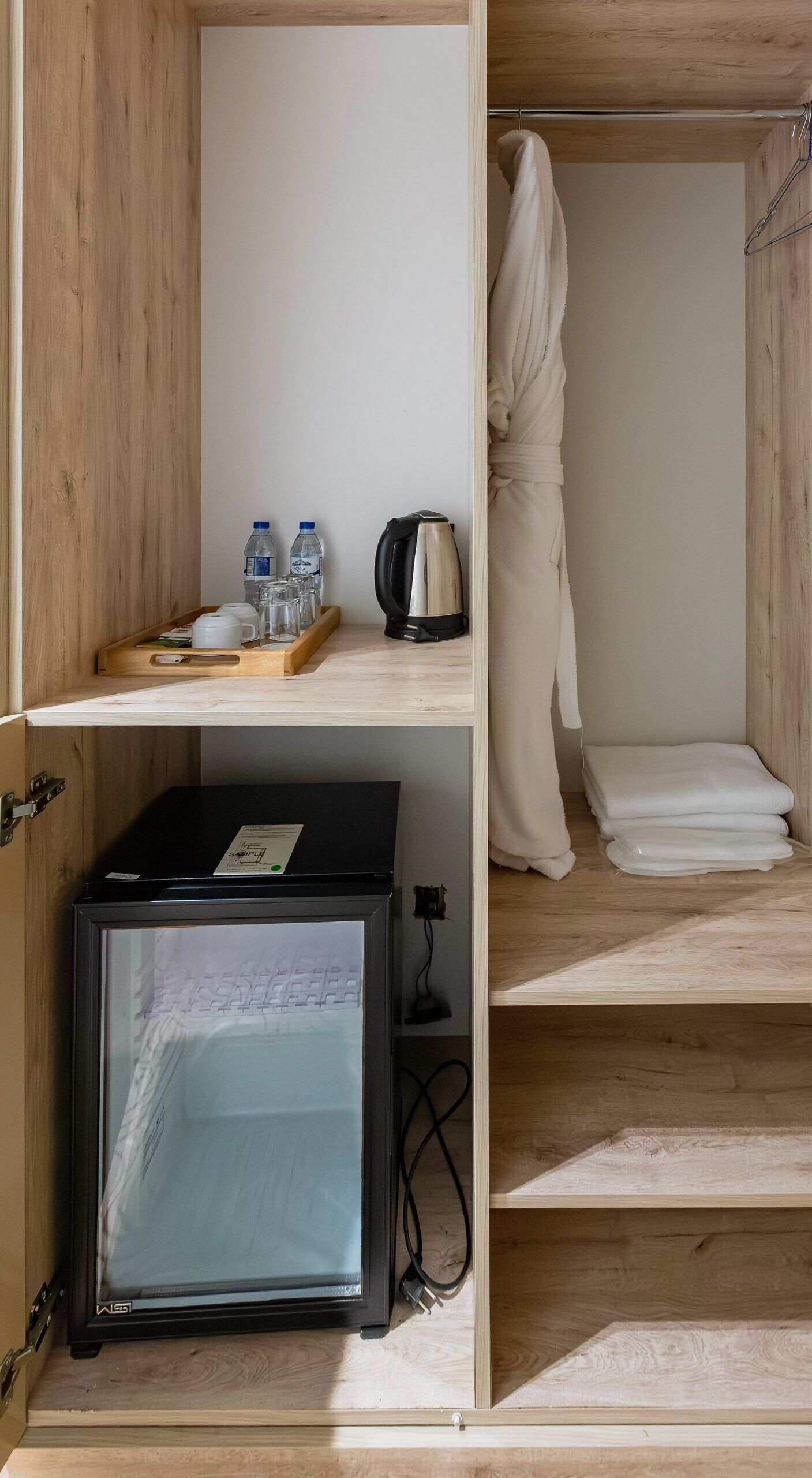 A compact room with a wooden desk, a refrigerator, a bed with a white blanket, and a robe hanging on the wall.
