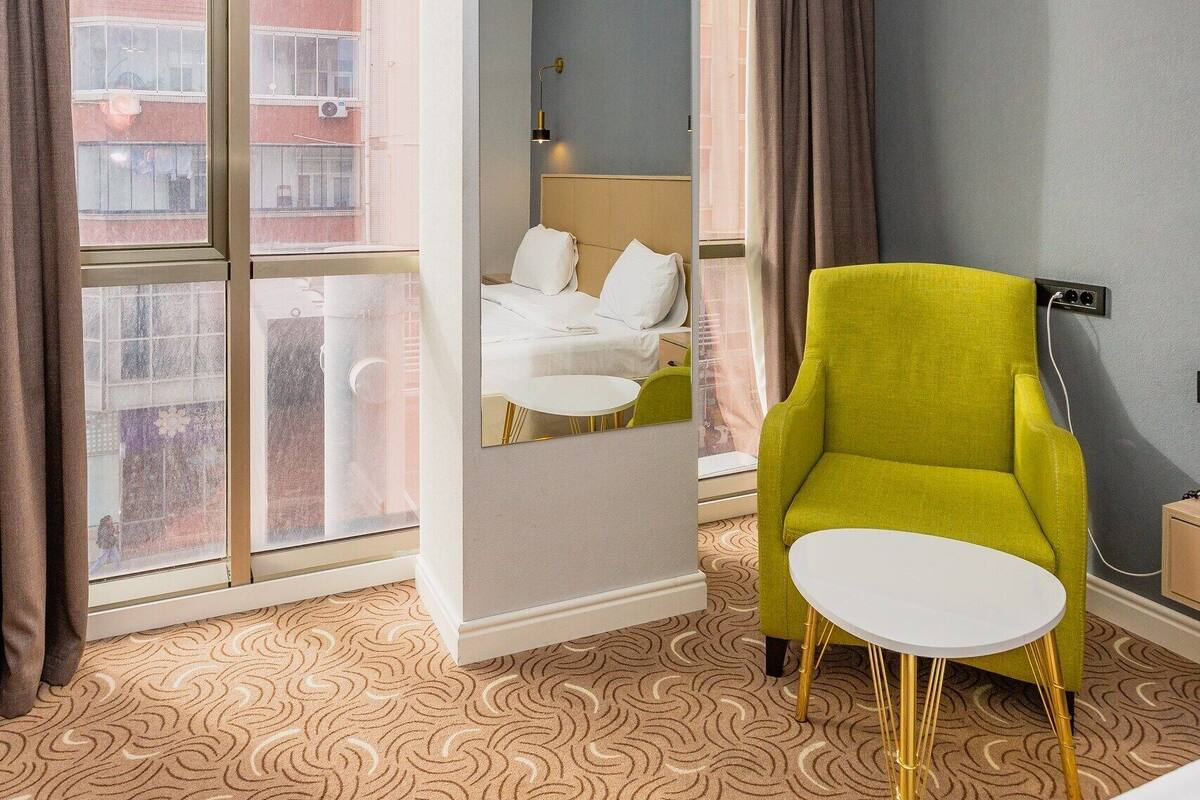 Ein Hotelzimmer mit einem Bett, einem grünen Sessel, einem kleinen weißen Tisch und einem großen Fenster mit Vorhängen.