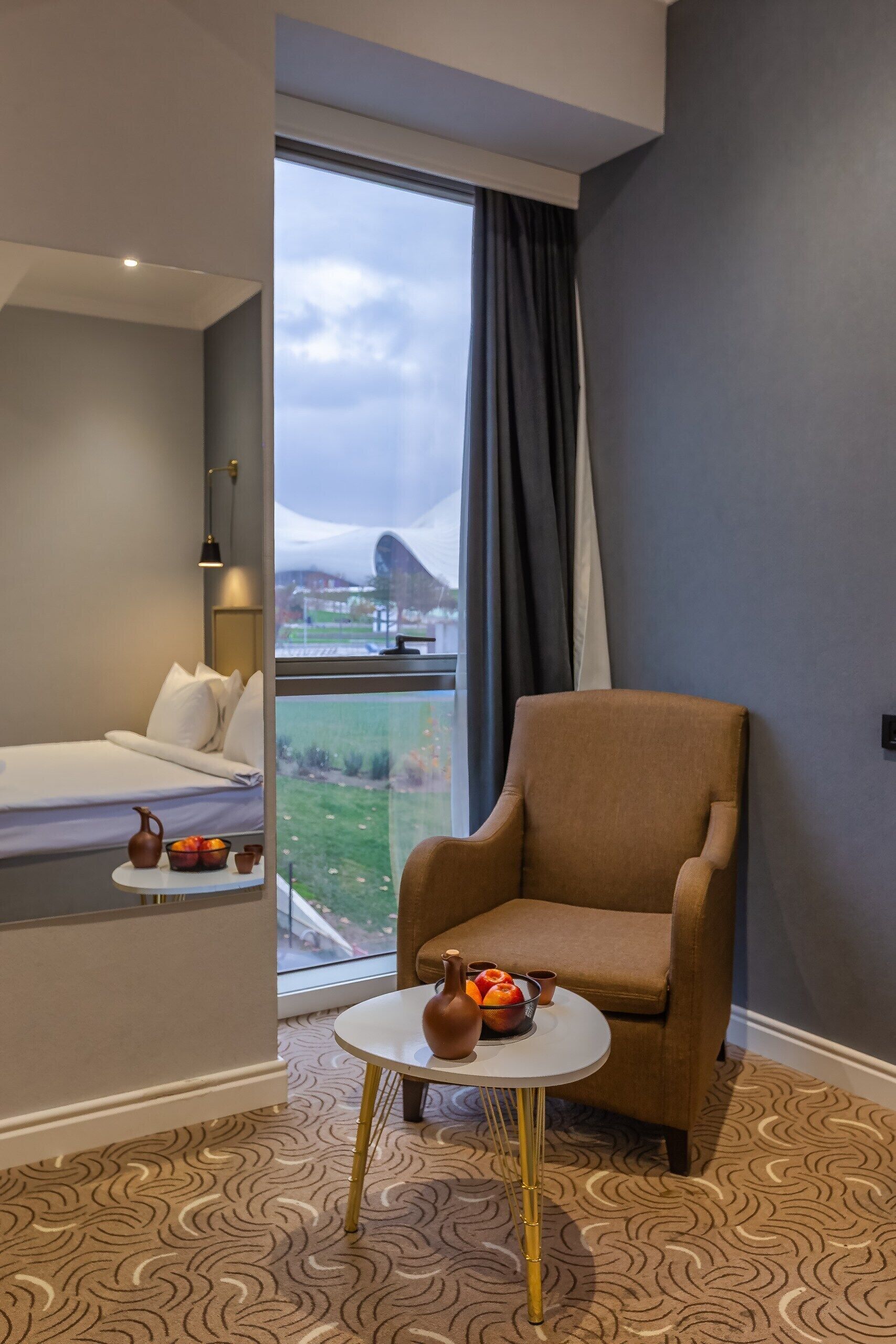 Una habitación de hotel moderna con un ventanal grande, una cama, un sillón marrón y una mesita con objetos decorativos.