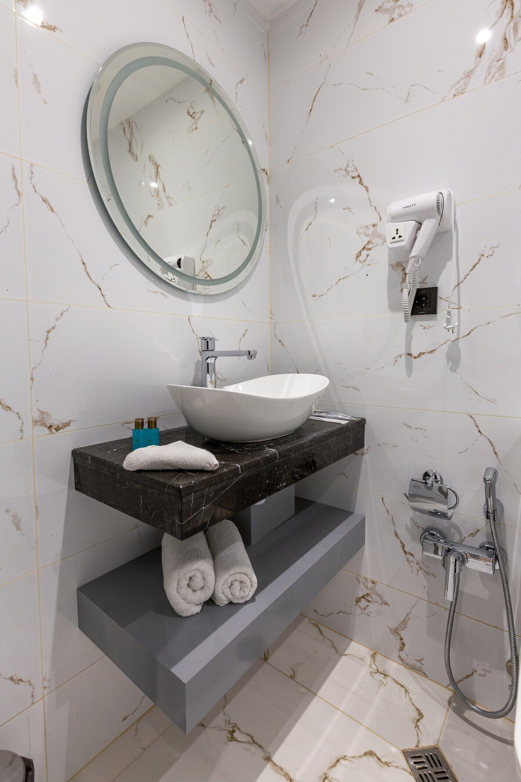 A bathroom with a marble-tiled floor, a white sink with a marble countertop, a round mirror, a hairdryer, and a shower.