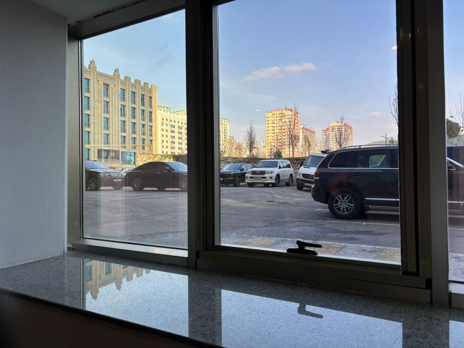 A view through a window of a parking lot with several cars and high-rise buildings in the background.