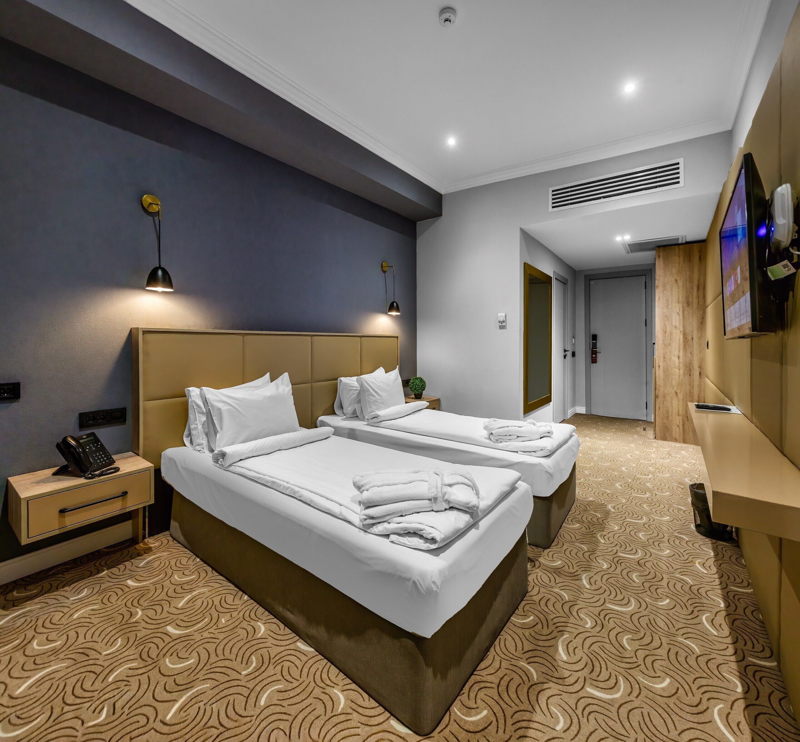 Quarto de hotel com duas camas, telefone ao lado da cama, uma mesinha e uma televisão fixada na parede.