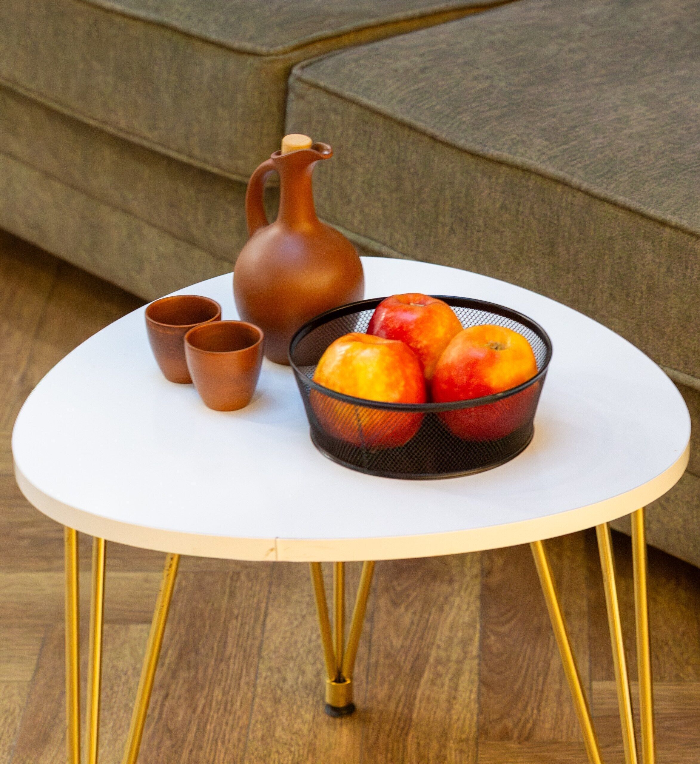 A small round table with a white top and gold legs, a black fruit basket with apples, a brown ceramic pitcher, and two brown cups on the table.