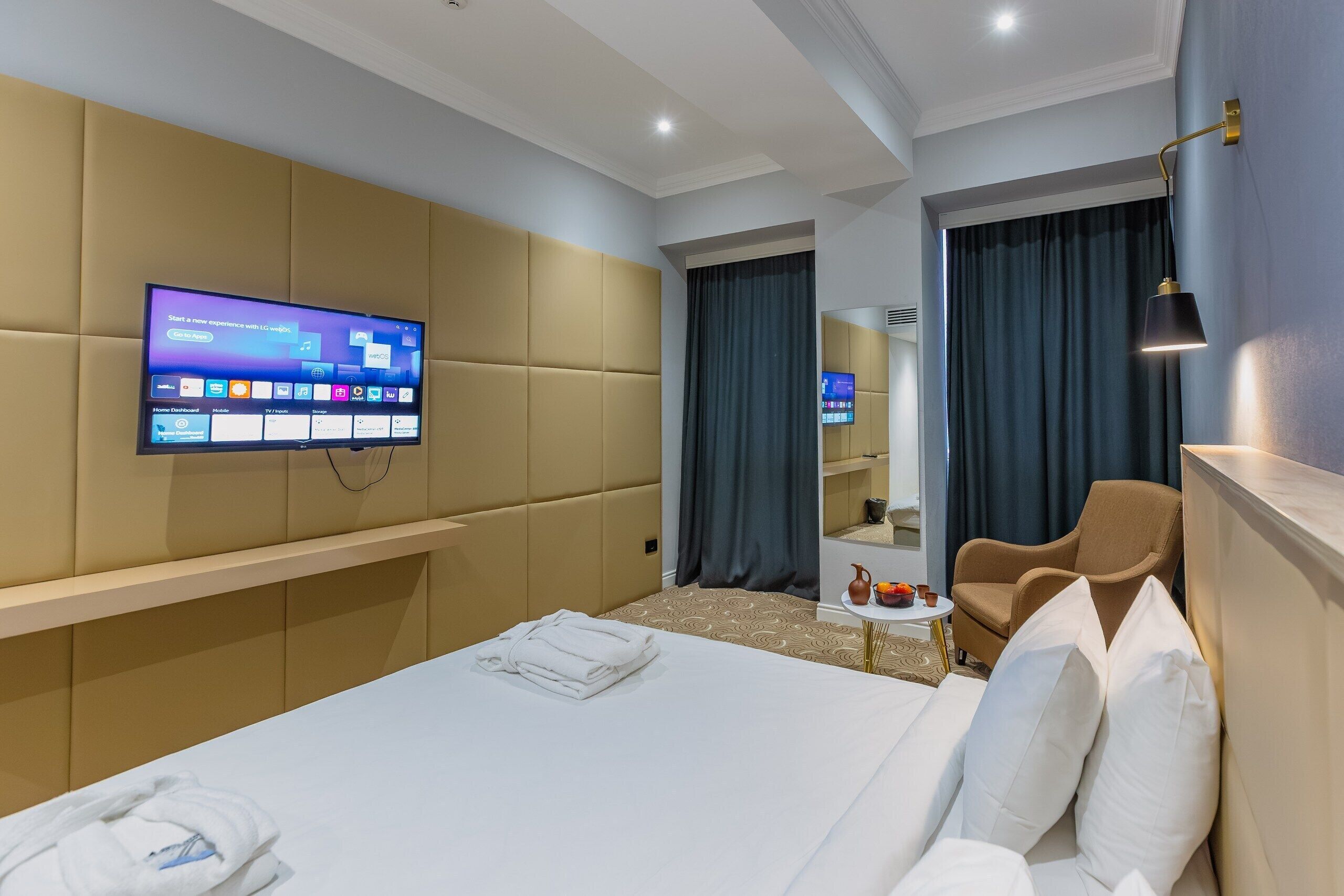 Une chambre d’hôtel moderne dotée d’un grand lit, d’un téléviseur à écran plat fixé au mur, d’un coin salon avec un fauteuil et une petite table, et d’un miroir au-dessus du lit.